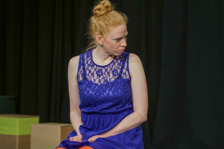Sitzende Frau mit roten Haaren, zum Dutt gebunden, ärmelloses lilanes Kleid, Bühnensituation, schaut skeptisch nach rechts.